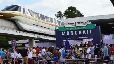 A crowd of people waits in line under a "Magic Kingdom Monorail" sign, while a gold-striped monorail train passes on an elevated track above the entrance. Trees and a station building are in the background.