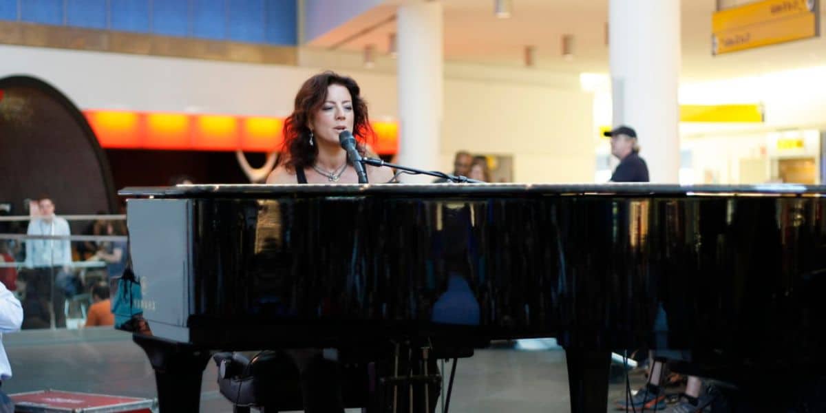 A woman with wavy dark hair sits at a black grand piano, singing into a microphone in a brightly lit indoor space with people and colorful lights in the background.