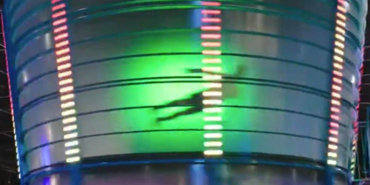A person is spinning inside a large, cylindrical, metallic structure with colorful vertical lights and a green glow, creating a blurred silhouette against the illuminated surface.