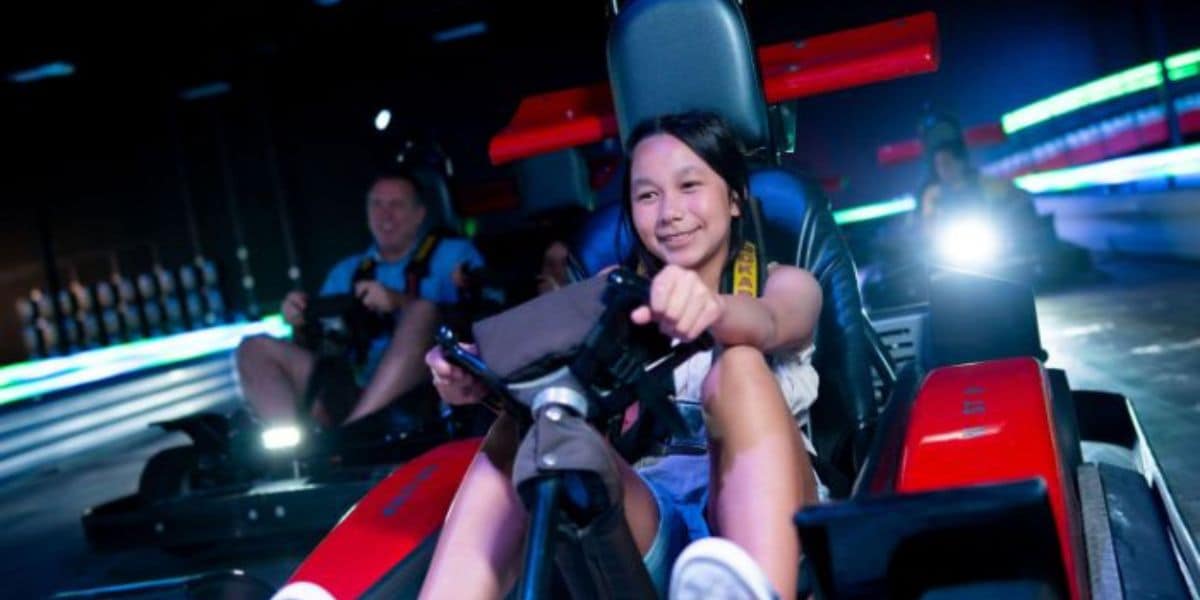 A smiling girl drives a red go-kart on an indoor track, with colorful lights and another racer visible in the background.