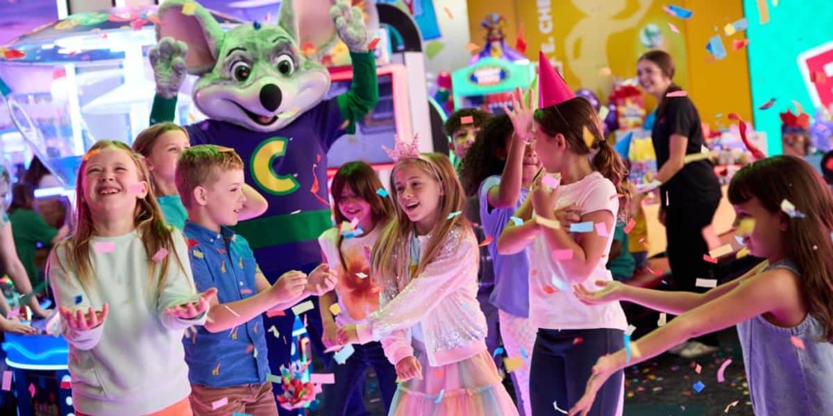 Children celebrate at an arcade as colorful confetti falls. A large mouse mascot in a green and purple outfit stands behind them, raising its arms. The kids are smiling, laughing, and wearing party hats. Bright arcade games fill the background.