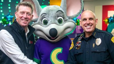 Two men, one in a black vest and one in a police uniform, stand on either side of a person in a Chuck E. Cheese mouse costume, all smiling for the camera indoors with colorful decorations in the background.