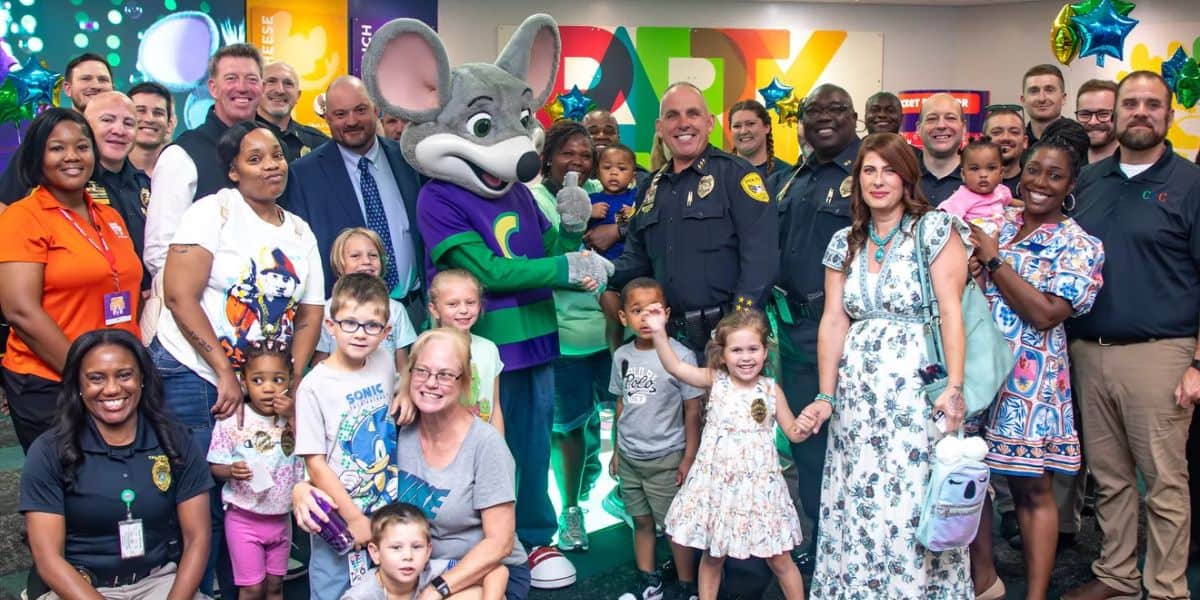 A large group of adults and children, including police officers and a person in a Chuck E. Cheese costume, pose and smile together at a brightly decorated party venue.