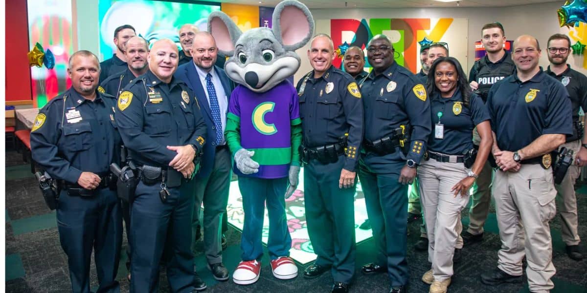 A group of smiling police officers poses indoors with a person in a Chuck E. Cheese mouse costume, standing in front of colorful party decorations and balloons.