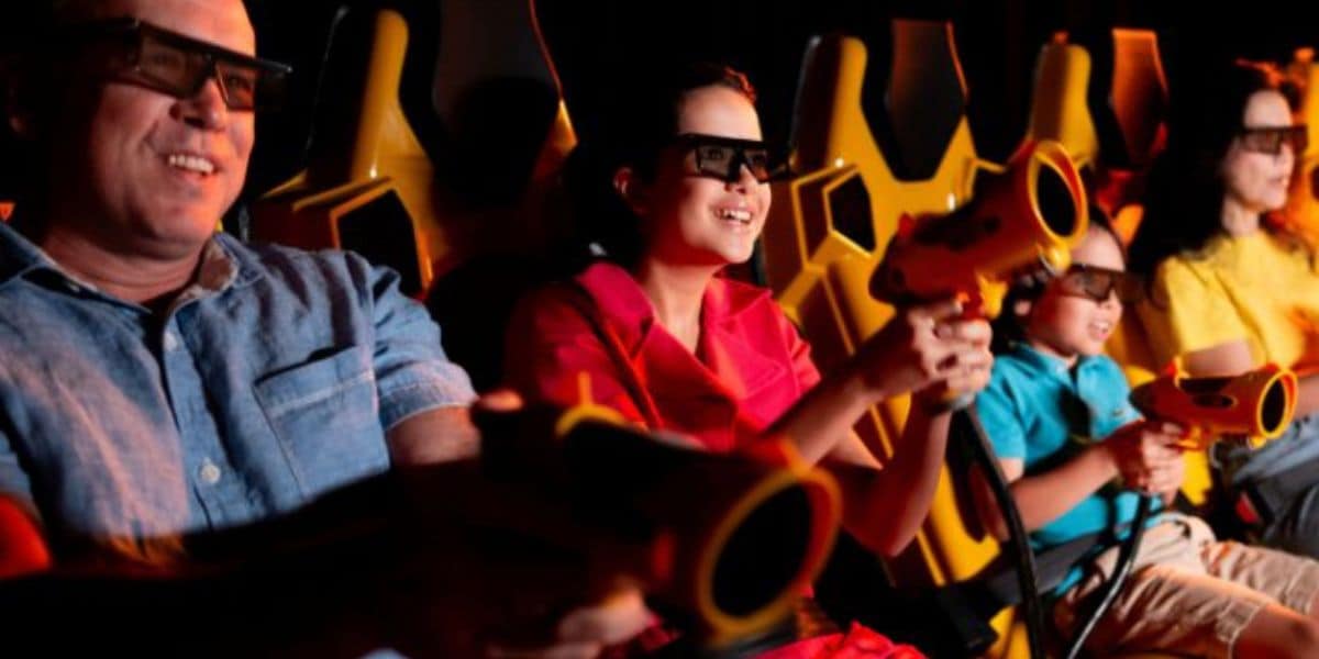 Four people wearing 3D glasses sit in yellow seats, holding toy guns and smiling, as if enjoying an interactive ride or game in a dimly lit entertainment venue.