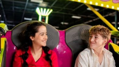 Two smiling teenagers sit side by side on a bright pink and yellow amusement park ride, looking at each other. Neon lights and arcade games are visible in the background.