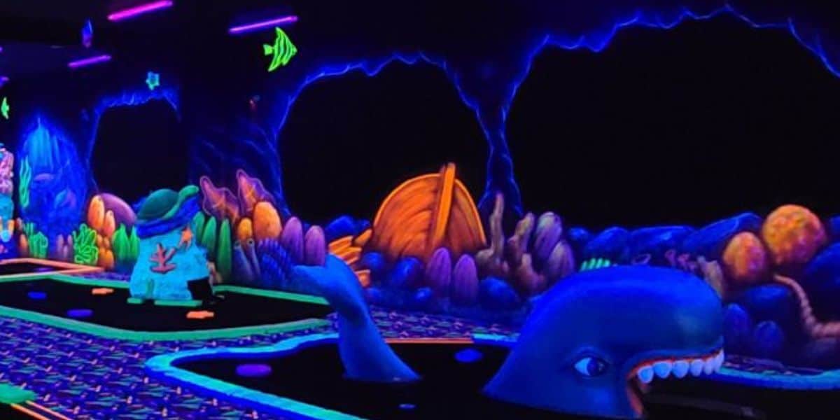 A glowing, underwater-themed mini-golf course with neon blue whales, colorful sea creatures, and vibrant coral decorations under blacklights.