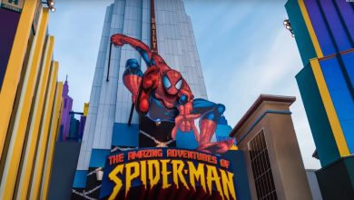 A colorful building facade features a large 3D Spider-Man figure above the entrance to "The Amazing Adventures of Spider-Man" ride at a theme park. The background shows stylized cityscape elements against a blue sky.