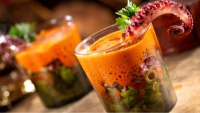 Two glasses filled with a layered salad of chopped vegetables, topped with a bright orange sauce and garnished with a cooked octopus tentacle and fresh herbs—a dish that looks straight out of a Disney fairytale.