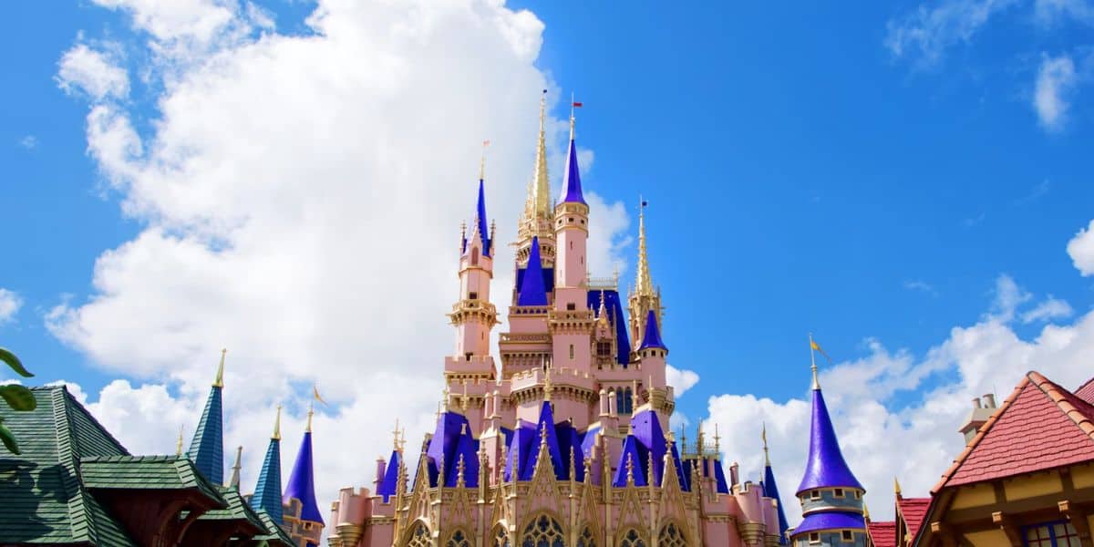 A bright blue sky with fluffy white clouds above a fairytale castle featuring pink walls and tall, pointed blue and gold spires, surrounded by rooftops.