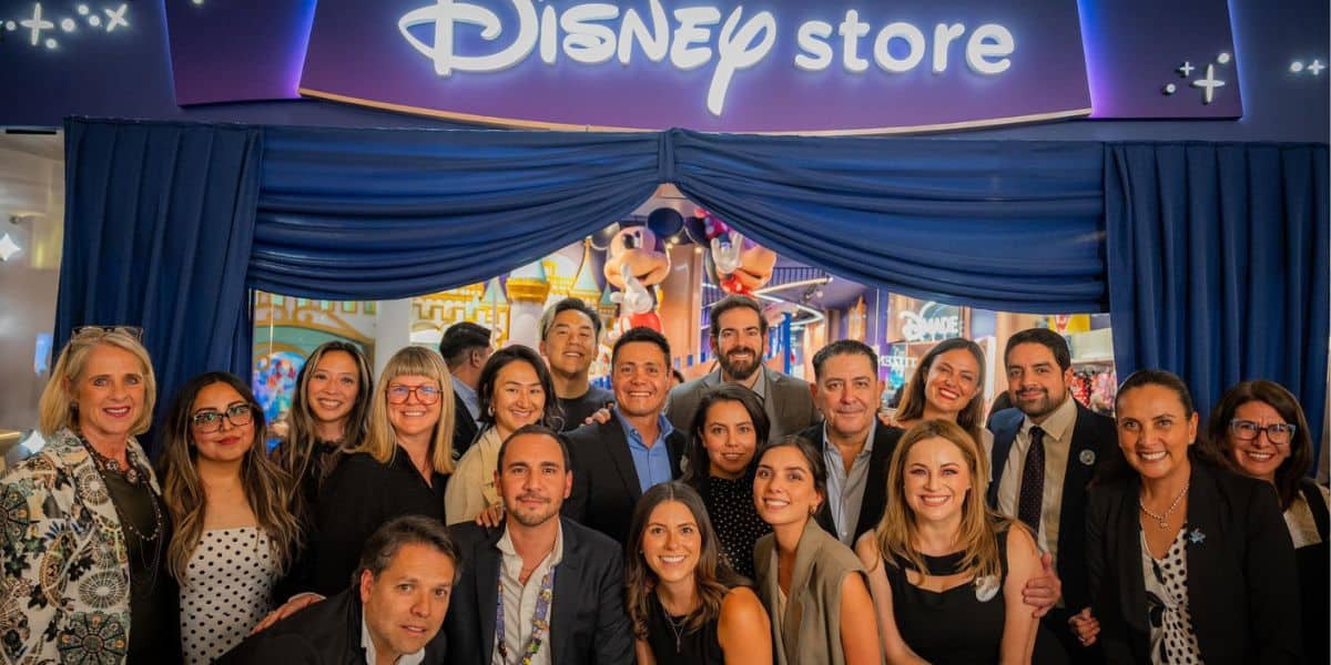 A group of people smiling and posing together in front of a Disney Store entrance with a large Disney logo above and Mickey and Minnie Mouse figures visible in the background.