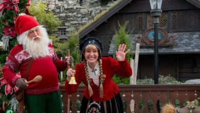 A man dressed as a Christmas gnome and a woman in festive Scandinavian costume stand by a decorated tree, smiling and waving, celebrating the joy of Christmas in front of a rustic wooden house at Disney World.