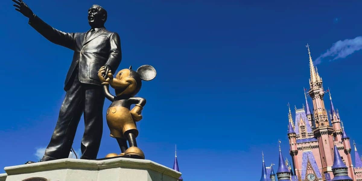 A bronze statue of a man and Mickey Mouse stands with the man pointing forward; behind them is a pink and blue castle against a clear blue sky.