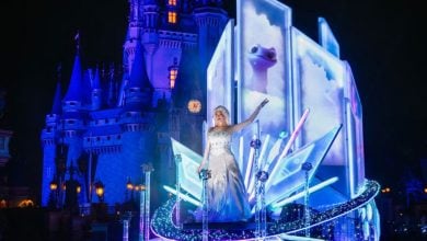 A woman dressed as Elsa from Frozen stands on a glowing icy-themed float, with a castle lit in blue in the background, dazzling spectators during a magical nighttime parade at a theme park.