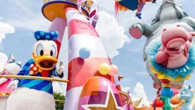 Donald Duck waves from a colorful parade float, surrounded by balloons and Disney-themed decorations, with Dumbo and other playful characters in the background under a bright, partly cloudy sky.