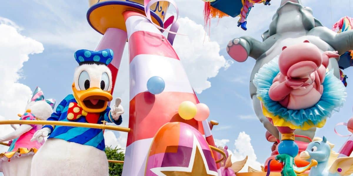 Donald Duck waves from a colorful parade float, surrounded by balloons and Disney-themed decorations, with Dumbo and other playful characters in the background under a bright, partly cloudy sky.