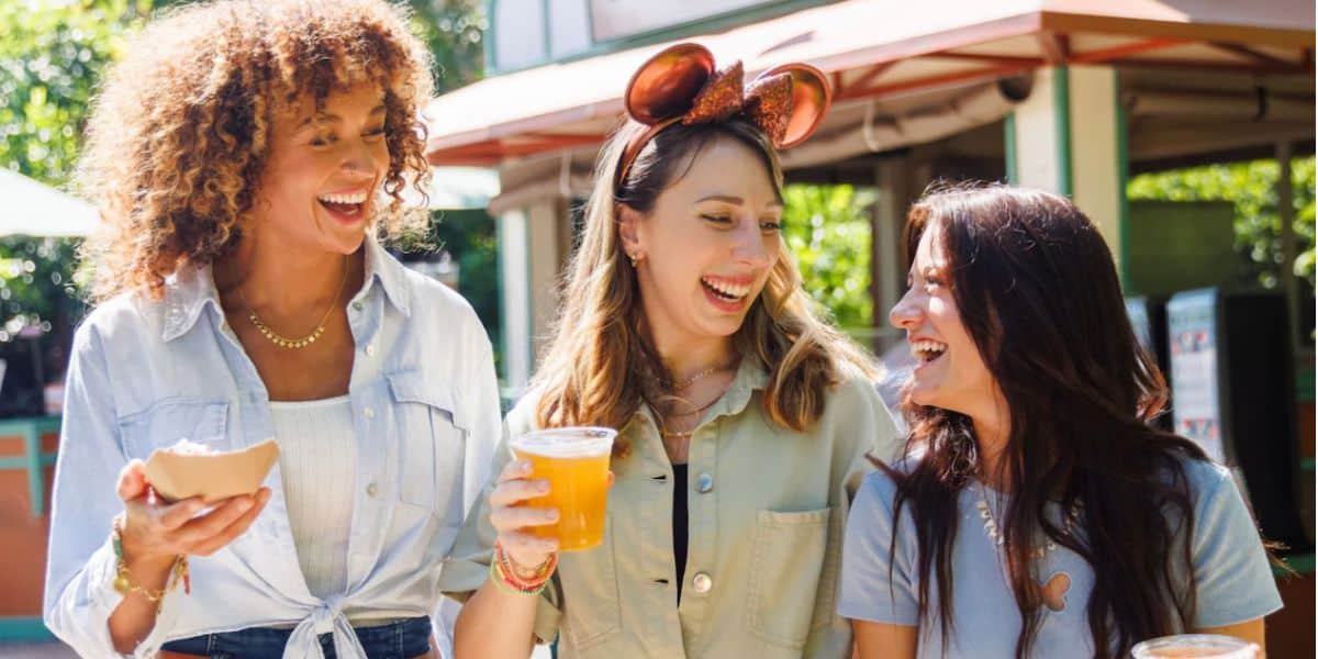 Three young women smiling and laughing outdoors at Epcot, one holding food, another with a drink, and the third wearing shiny mouse-ear headbands. They appear to be enjoying a sunny day together.