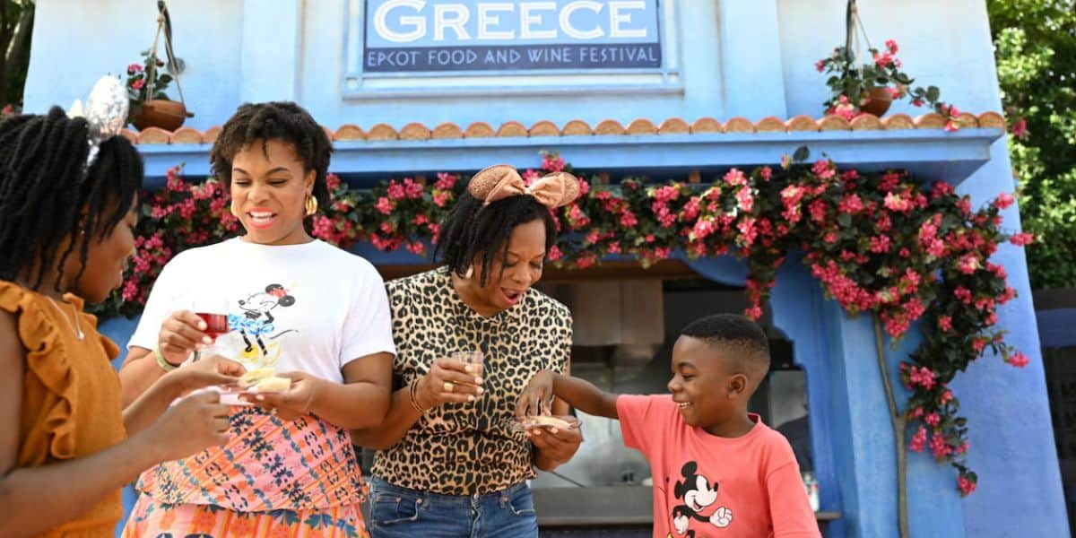 A family of four smiles and enjoys food in front of a booth decorated with flowers and a sign reading “Greece: EPCOT Food and Wine Festival.” They wear casual clothes, some with Mickey Mouse designs, celebrating the flavors of EPCOT together.