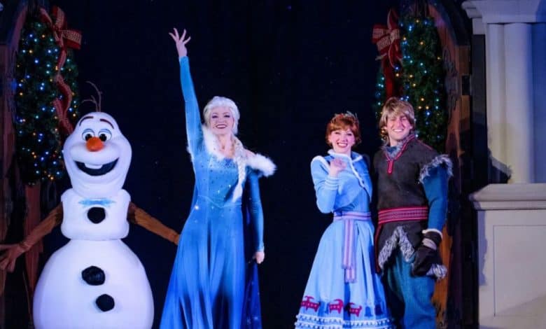 Three performers dressed as Olaf, Elsa, and Anna from "Frozen" and another as Kristoff stand on stage, smiling and waving amid christmas decorations and sparkling Christmas trees in the background.