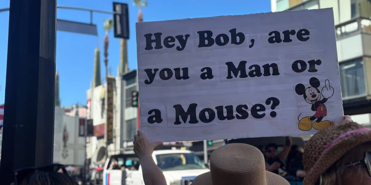 A person holds a sign in a city street that reads, "Hey Bob, are you a Man or a Mouse?" with an image of Mickey Mouse standing next to the text.