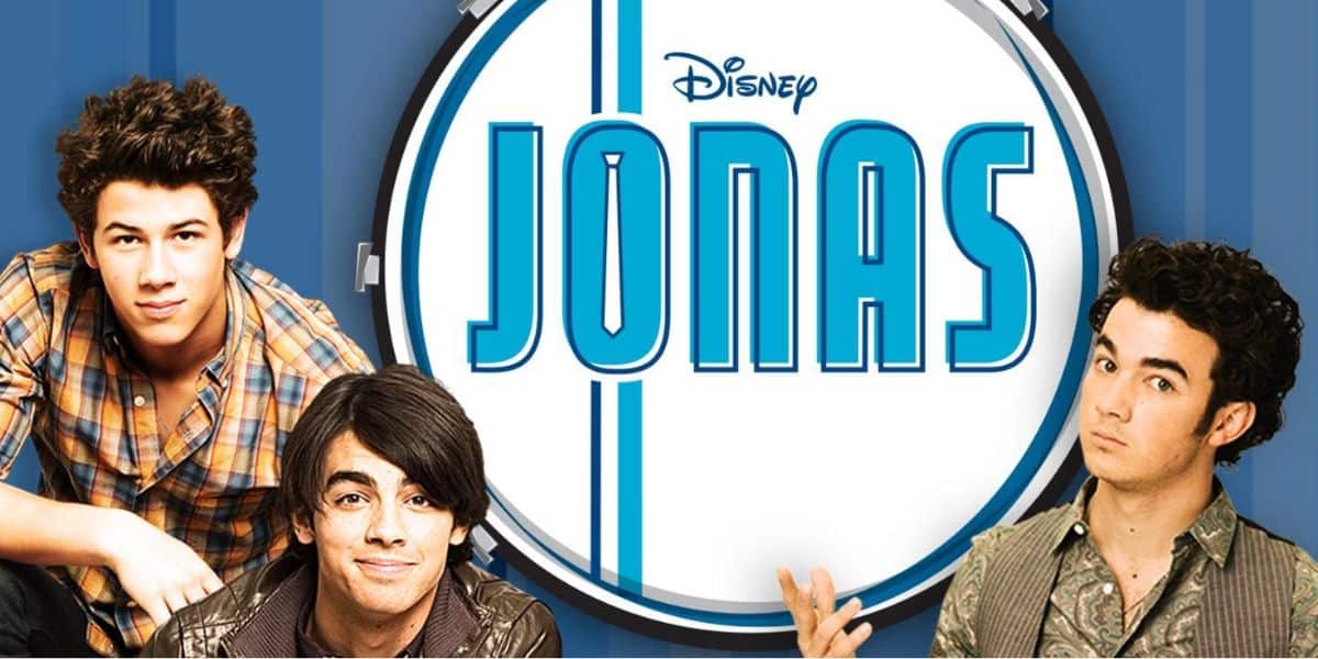 Three young men, the Jonas Brothers, pose in front of a large drum with the Disney “JONAS” logo. The blue background with white stripes adds energy as they smile in their casual clothes.