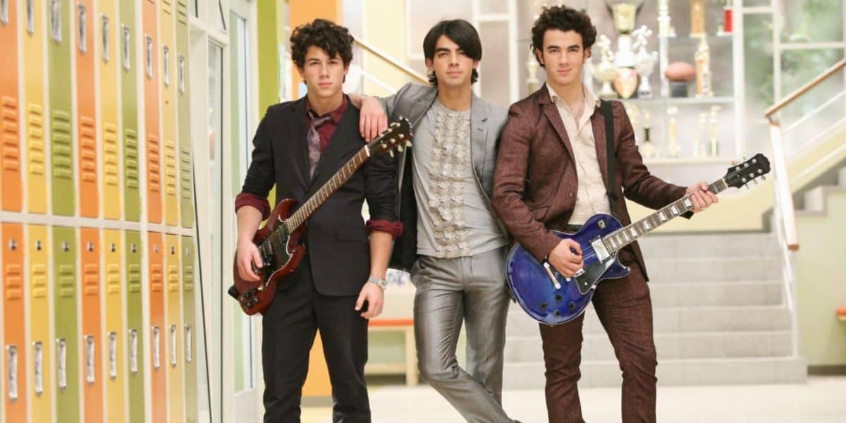 Three young men, dressed in suits like the Jonas Brothers, stand in a school hallway lined with yellow lockers; two hold electric guitars while trophies glimmer on shelves behind them.