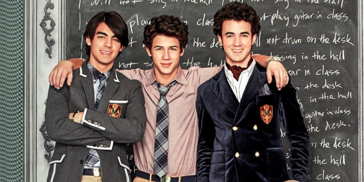 Three young men with dark, curly hair wearing school uniforms stand smiling in front of a blackboard, reminiscent of the Jonas Brothers. They have their arms around each other and appear happy and relaxed.