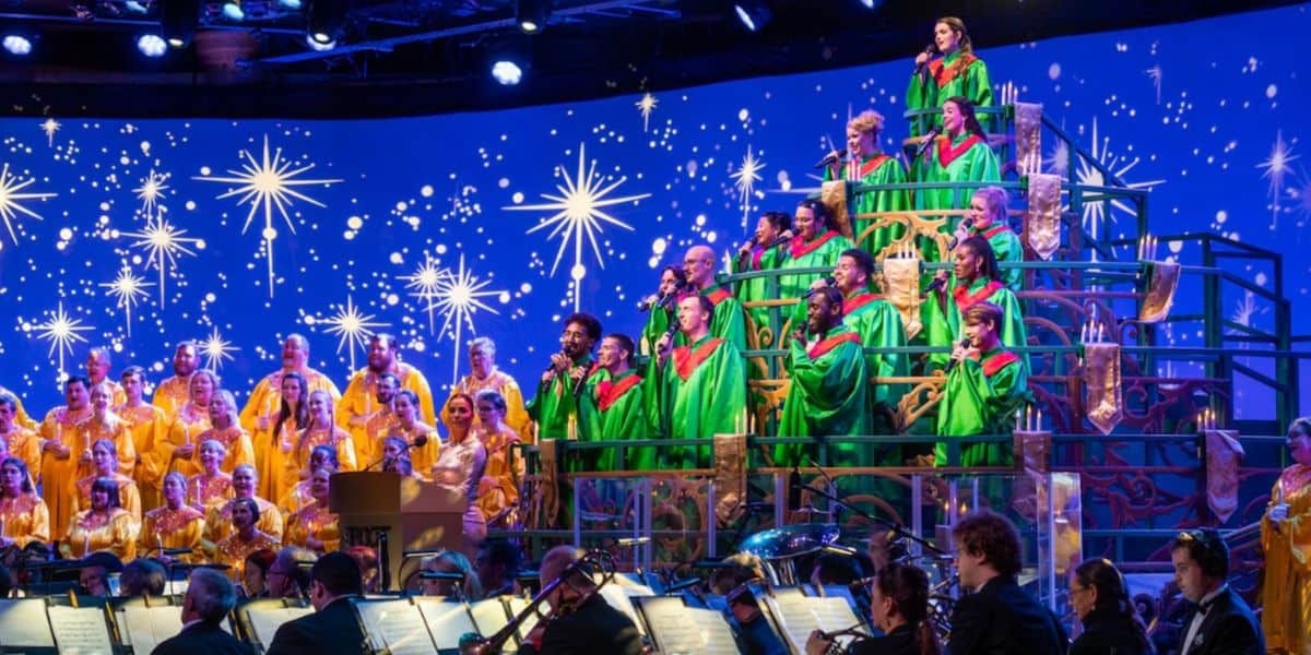 A large choir dressed in gold and green robes sings on tiered stands, with an orchestra playing in front. The background features a vibrant blue screen with bright, glowing stars, creating a festive Christmas atmosphere.