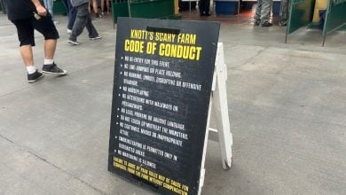 A sandwich board sign on concrete displays the "Knott's Scary Farm Code of Conduct," listing rules such as no fighting, no inappropriate language, and no disruptive behavior. People are walking nearby.