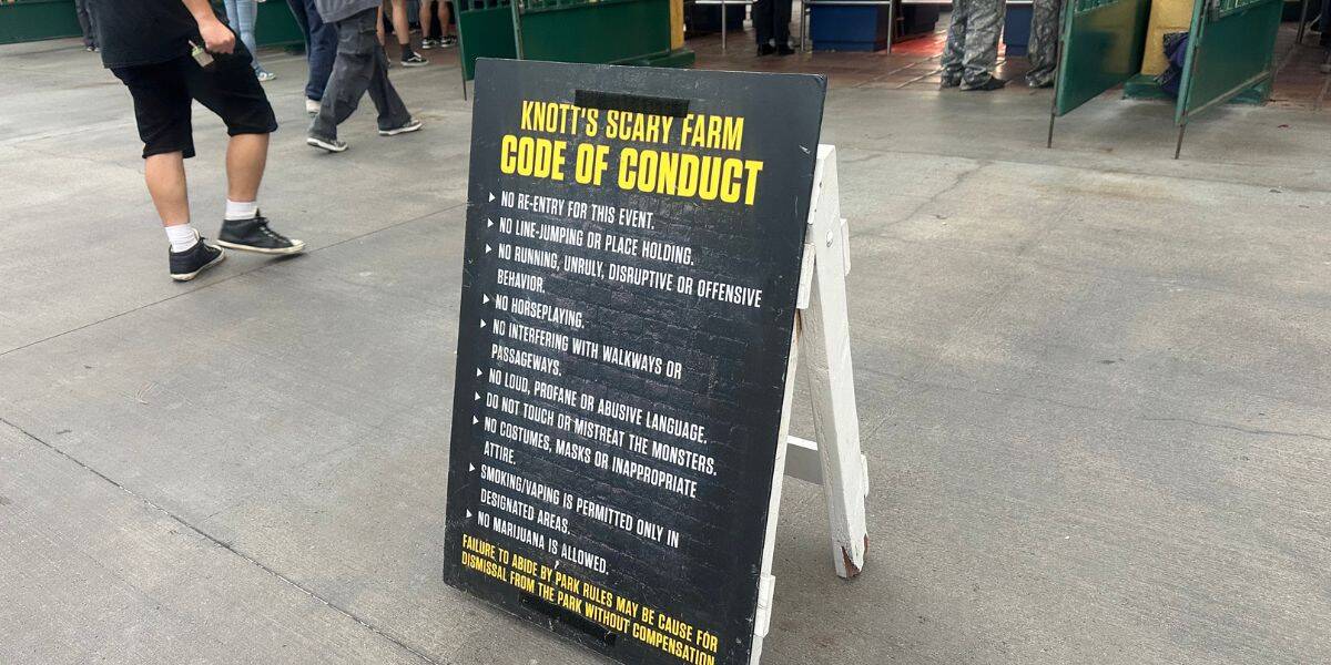 A sandwich board sign on concrete displays the "Knott's Scary Farm Code of Conduct," listing rules such as no fighting, no inappropriate language, and no disruptive behavior. People are walking nearby.