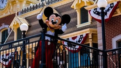 Mickey Mouse stands on a balcony decorated with red, white, and blue banners, giving two thumbs up in front of a building with a clock and ornate details.