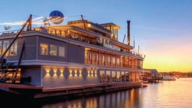 A large, multi-level riverboat restaurant with string lights is docked by the water at sunset, evoking a Disney-like charm as colorful shades of orange and blue reflect across the calm water.