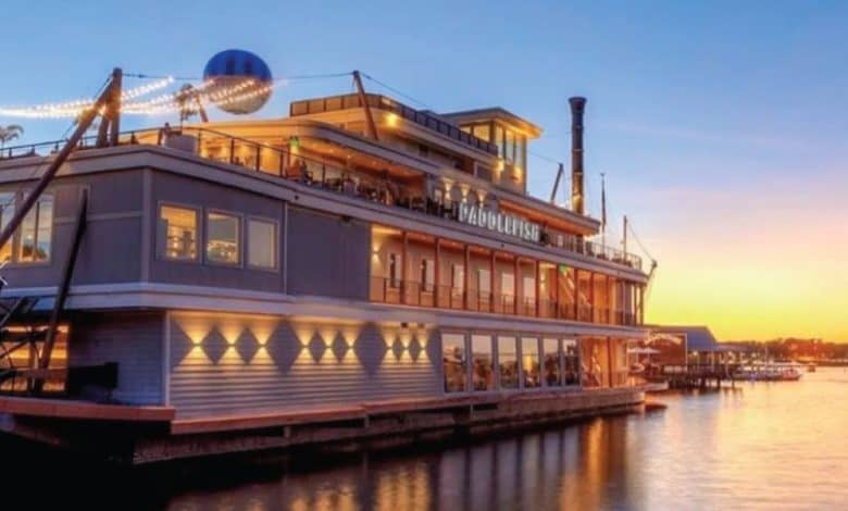 A large, multi-level riverboat restaurant with string lights is docked by the water at sunset, evoking a Disney-like charm as colorful shades of orange and blue reflect across the calm water.