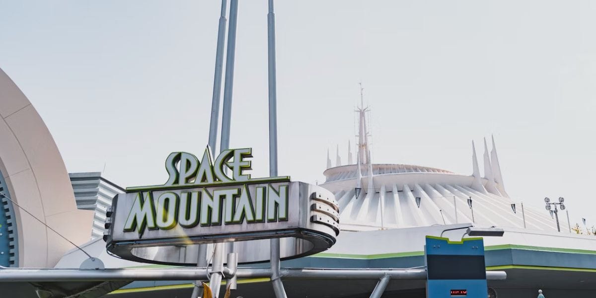 The Space Mountain sign stands in front of the futuristic, white, spiked dome of the Space Mountain ride at Disneyland, under a clear sky.