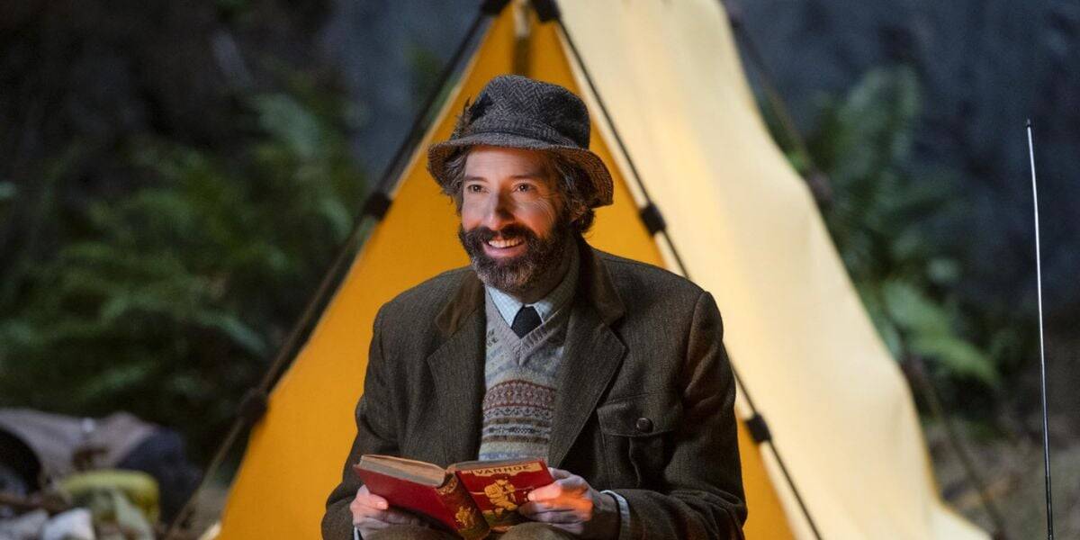 A bearded man in a hat and tweed jacket smiles while sitting in front of a yellow tent, holding an open book, with greenery in the background—capturing a scene as charming as a classic Disney adventure.