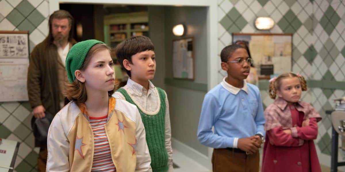 Four children stand closely together indoors, looking attentively ahead; a man stands in the background near a doorway, watching them. The green and white checkered walls and vintage feel create a scene reminiscent of classic Disney storytelling.