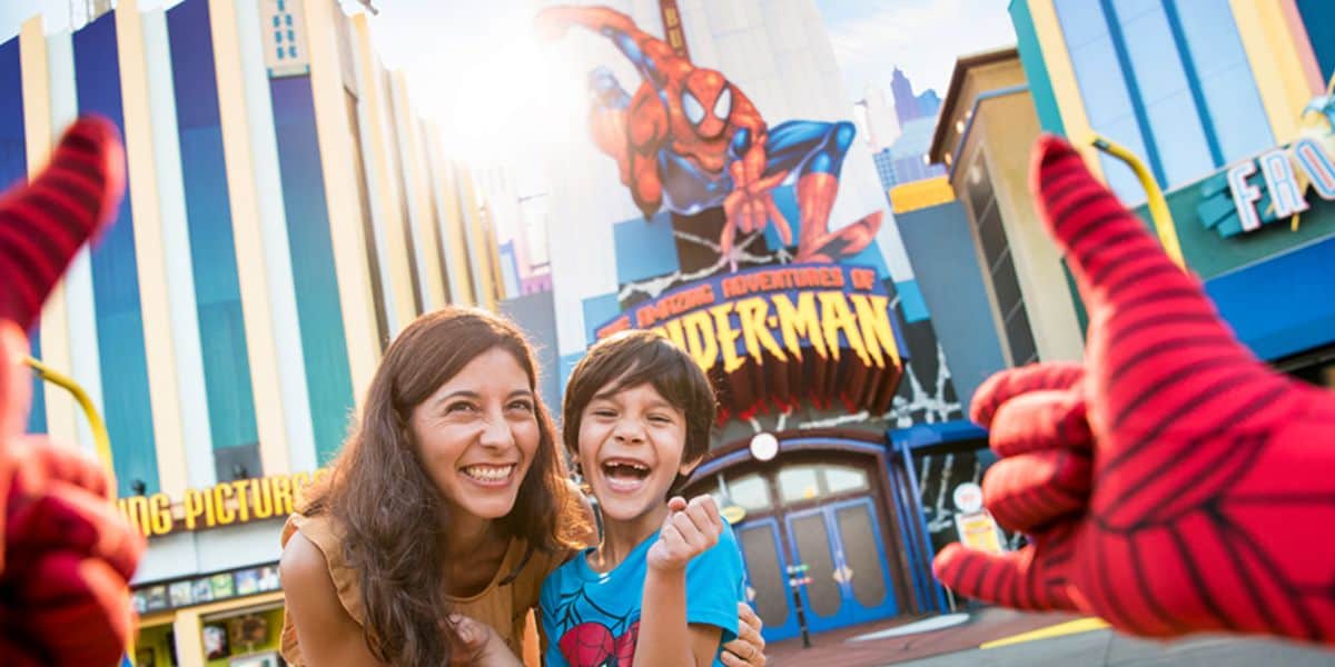 A smiling woman and boy pose in front of the spider-man ride at a theme park. Both look excited, while two gloved hands mimic Spider-Man’s web-shooting gesture in the foreground.