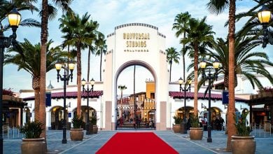 Entrance to Universal Studios Hollywood, featuring a large white arch with the park’s name, palm trees on both sides, vintage-style street lamps, and a red carpet leading to the gate.