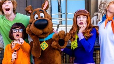 Five people dressed as Scooby-Doo characters—Shaggy, Velma, Scooby, Daphne, and Fred—stand together making surprised faces in front of a building with statues, bringing the classic scooby gang to life.