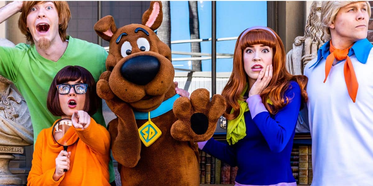 Five people dressed as Scooby-Doo characters—Shaggy, Velma, Scooby, Daphne, and Fred—stand together making surprised faces in front of a building with statues, bringing the classic scooby gang to life.