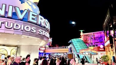 Crowds stroll under bright lights at the Universal Studios Store at night. Colorful neon signs and universal-themed buildings light up the street beneath a dark sky, with the moon shining above.