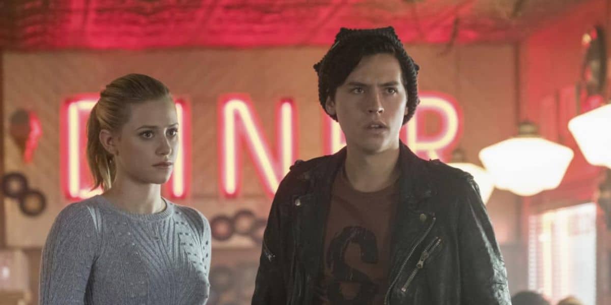 Two young people stand inside a retro-style Riverdale diner bathed in red neon lights. The woman has blonde hair pulled back and a light sweater, while the man wears a dark beanie and leather jacket. Both look concerned.