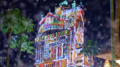 The Hollywood Tower Hotel building is brightly lit with colorful holiday projections, including gingerbread men, with palm trees and artificial snow falling at night.