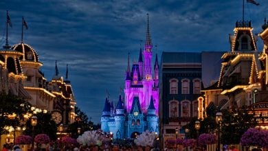 A vibrant night scene at a theme park, featuring a brightly lit fairy tale castle glowing in purple and blue, surrounded by ornate buildings with warm lights and a crowd of visitors on the main street.