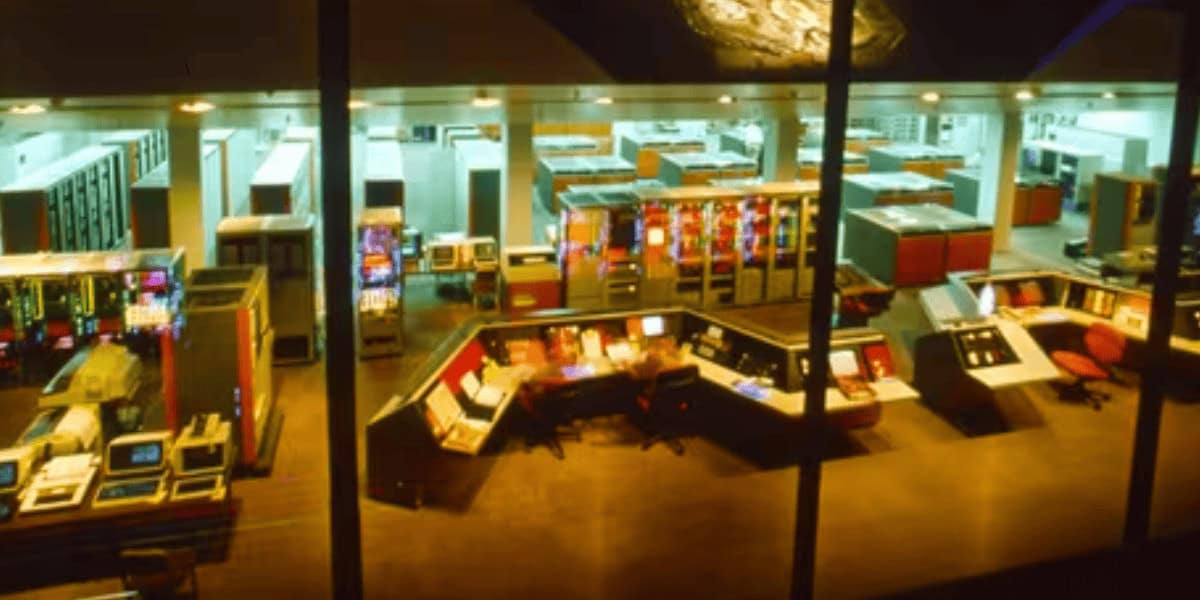 A brightly lit room filled with large, vintage computer equipment and control panels, with several workstations and monitors arranged in rows, viewed through a glass window.