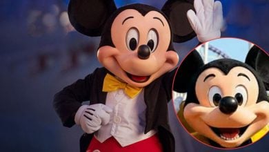 Mickey Mouse mascot in a black jacket, yellow bow tie, and red shorts waves with one hand. In the corner, a circular inset shows a close-up of Mickey Mouse’s smiling face.