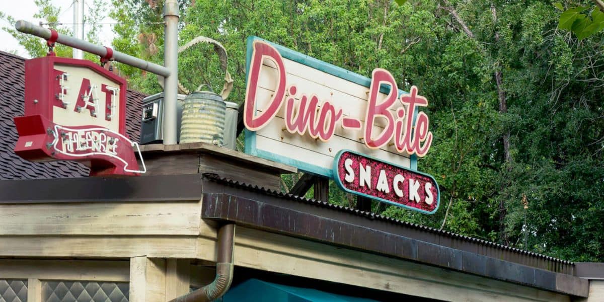 A retro-style sign reads "Dino-Bite Snacks" above a building at Disney, with a smaller red sign next to it that says "Eat Here." Green trees are visible in the background.