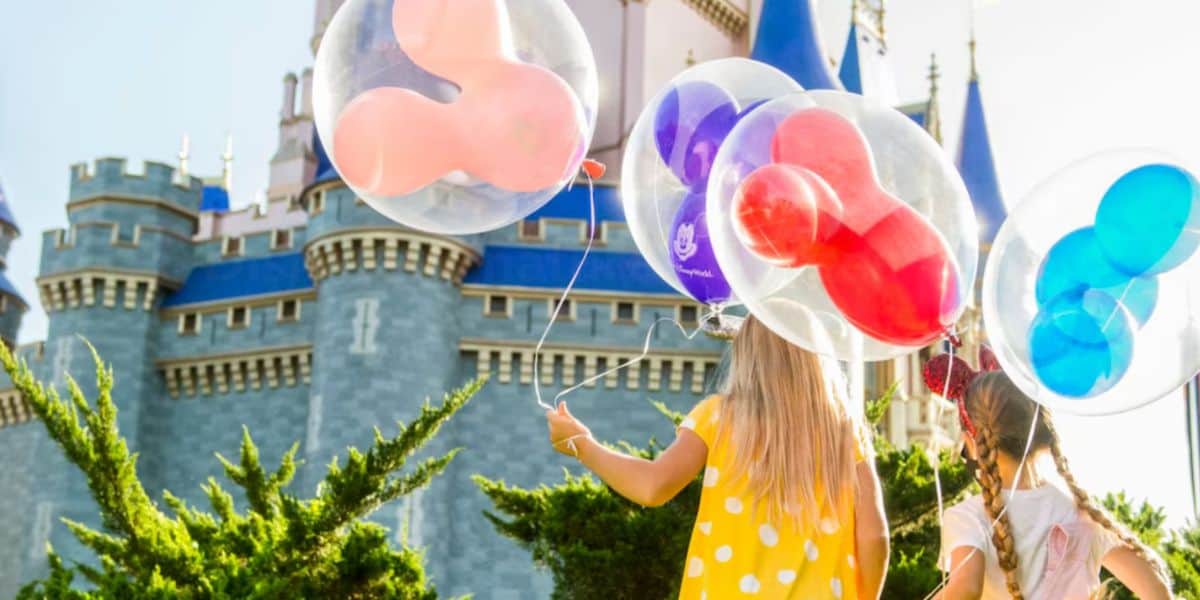 Two children holding colorful Mickey Mouse-shaped balloons stand in front of a fairytale Disney castle with blue spires on a sunny day. The children wear yellow and white polka-dotted and pink dresses.