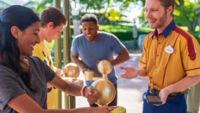 A smiling woman scans her Disney wristband at an entrance while a theme park employee in uniform assists her. Two other people wait in line behind her, also smiling, outside on a sunny day.