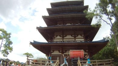 A group of performers in blue outfits play a large red taiko drum in front of a traditional five-story Japanese pagoda, capturing the vibrant spirit of Japan amidst trees under a partly cloudy sky.